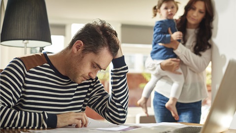 Homem com ar angustiado, em grande plano, e mulher com criança ao colo, em segundo plano. Ambiente doméstico.